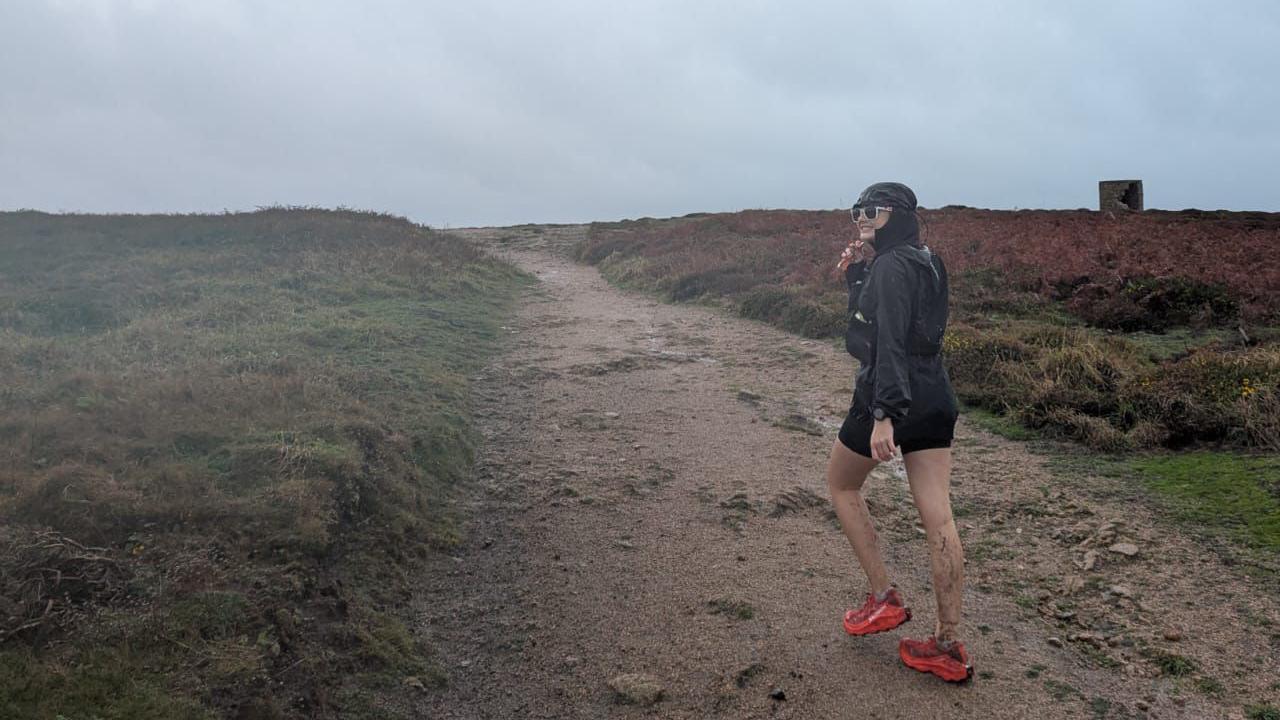 Emily is wearing a black jacket, shorts, and bright red trail shoes walking on a muddy path through open moorland. The sky is overcast with grey clouds, and the landscape features grassy areas and reddish-brown vegetation.
