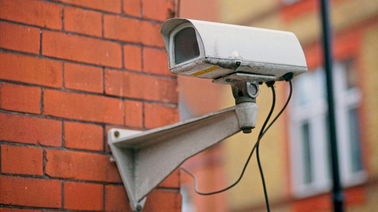 A white, rectangular, CCTV camera mounted on a red brick wall