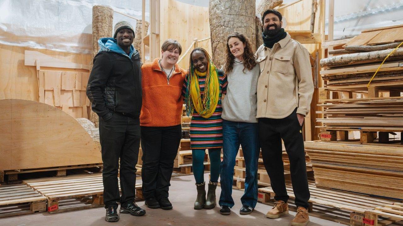 A group of five people - two men and three women - stand inside a room which has many wooden pallets in it, some on top of each other. The group are smiling at the camera and linking arms.