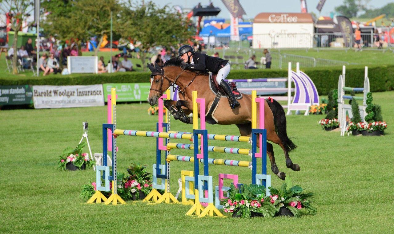 A horse rider dressed in black and white riding gear is in mid-action jumping over a colourful fence on the back on their brown horse. You can see several stalls in the distance and people sat on grass.