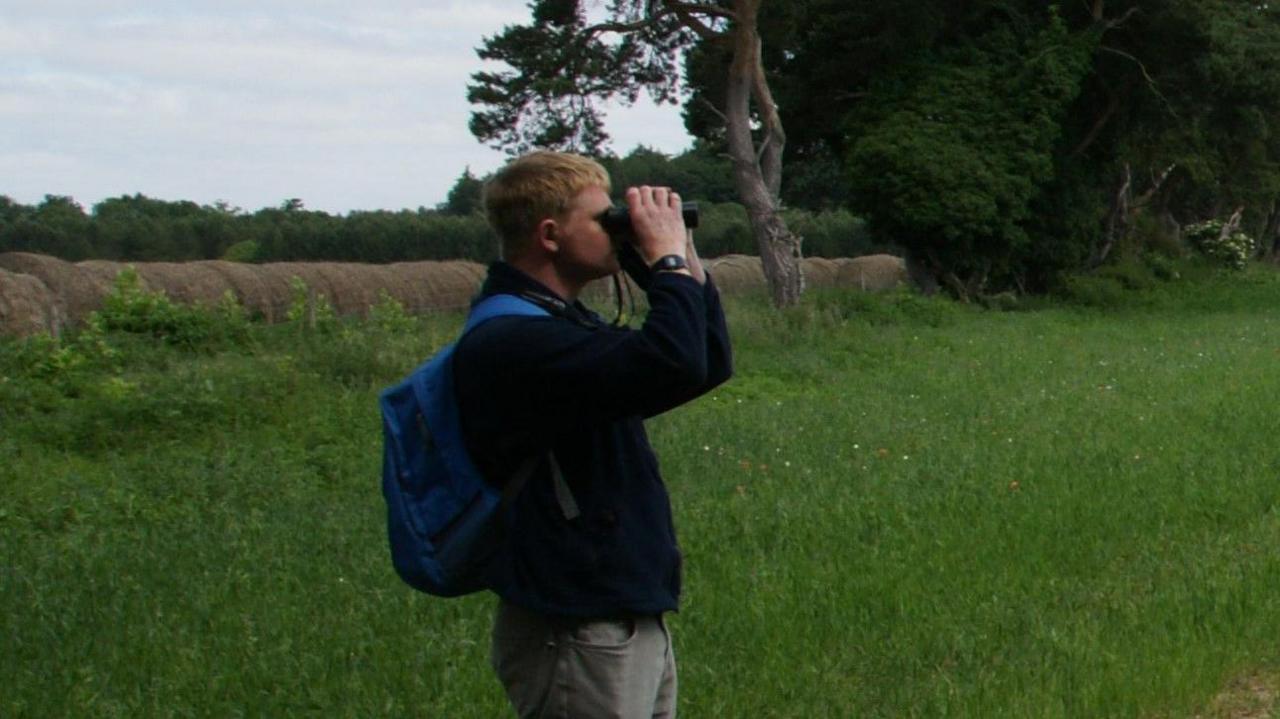 Tim Cowan is pictured in a grass field looking through binoculars for stone-curlew birds. He has short blond hair and is wearing a black jumper with grey trousers. He has a blue backpack on.