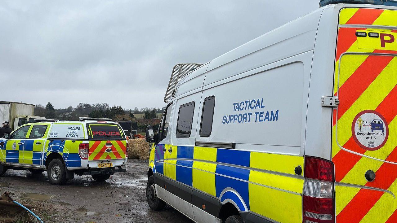 Two police vehicles on a field. The rear one is a van, marked "Tactical Support Team".