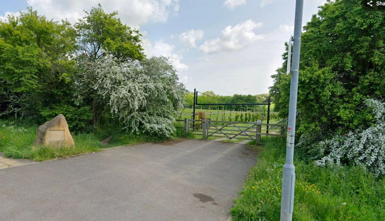 Entrance to a burial ground, showing black metal height restrictor and wooden farm gate with grass and young trees beyond. There is a road in the foreground, and a brown stone to the left has the words BURIAL GROUND carved into it. There are trees either side of the entrance .