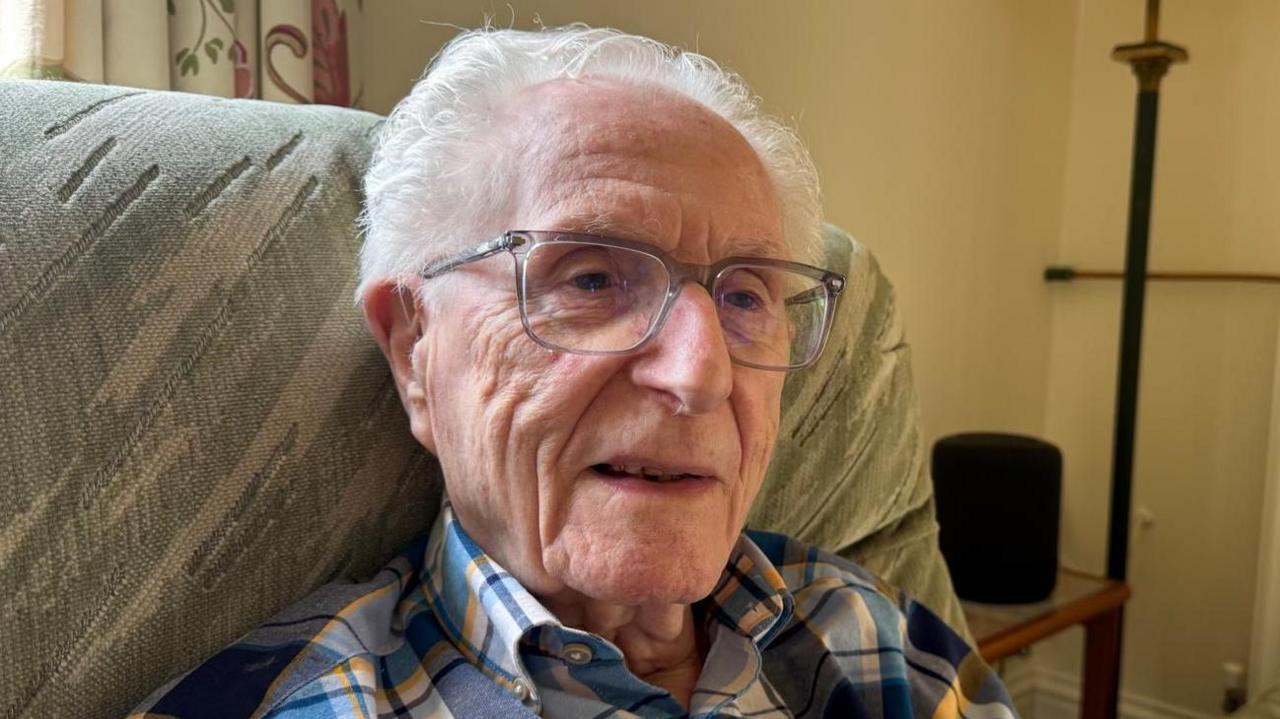A man sitting in a chair wearing glasses and a check shirt, looking slightly to the side of the camera.