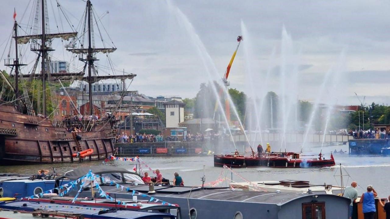 The ship Pyronaut fires water into the air from multiple points as it sits in the middle of the harbourside as part of the Bristol Harbour Festival. People are watching from other boats and also from the dockside.
