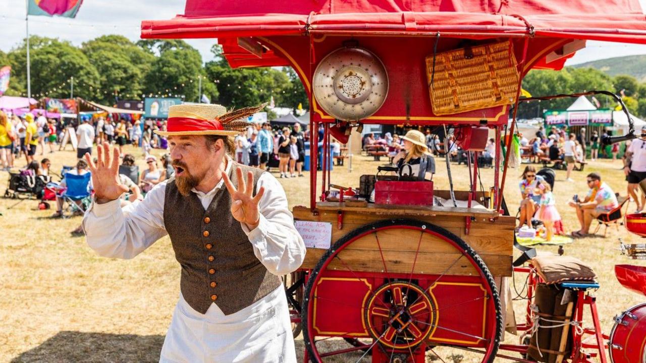 A performer dressed as a Victorian street trader stands in front of a red wooden cart with large wheels which has various things attached to its sides including a picnic basket and a large metal colander. He is wearing a straw boater and a waistcoat and is holding up nine fingers and thumbs.