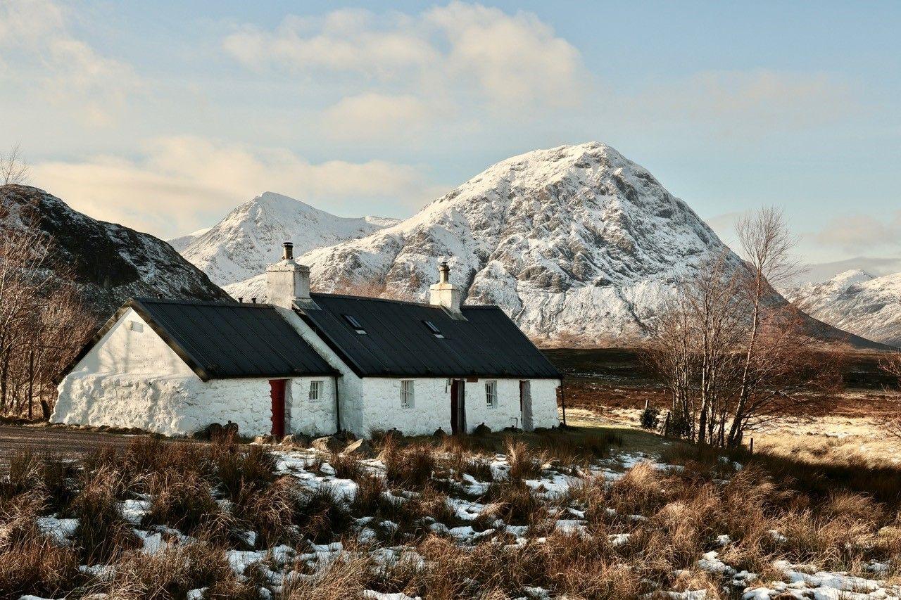 A small white cottage with a dark roof sits on frosty ground in front of a snow covered mountain peak