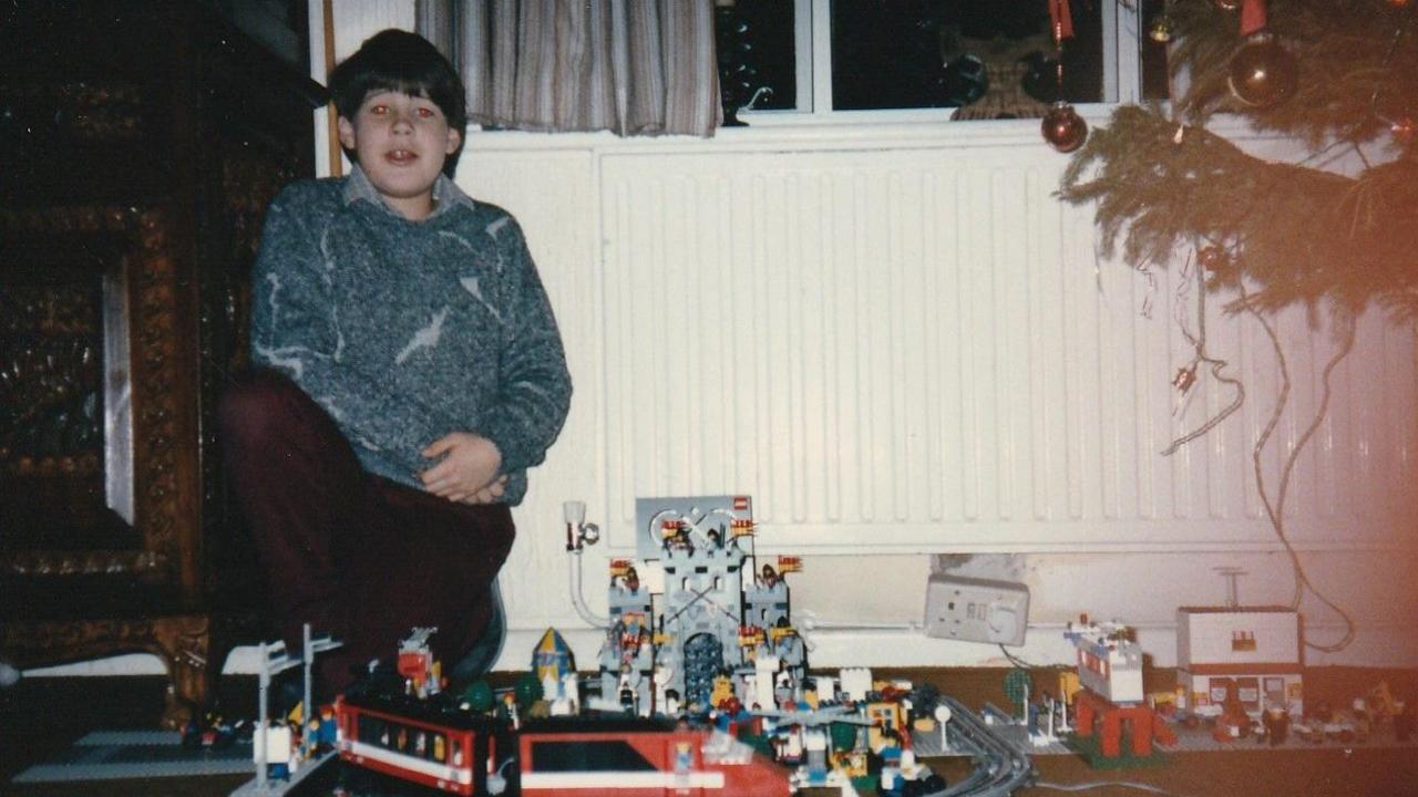 A childhood photo of Simon Papouis wearing a blue jumper crouched next to a Lego train set. He is to the left of a Christmas tree, and is in front of a white radiator.