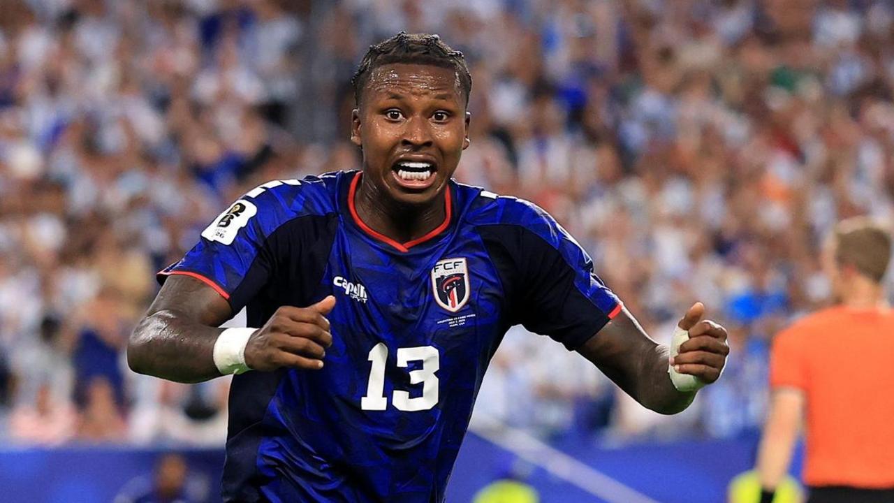 Sidny Lopes Cabral, visible from waist up wearing a dark blue Cape Verde shirt with a white number 13 on the chest, holds out his two hands in front of him with a suprised look of glee on his face as he celebrates a goal at the Fifa World Cup. Out of focus in the background are a referee wearing an orange shirt and a stand full of spectators