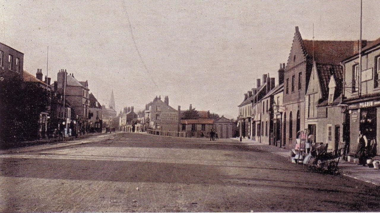 Black and white photo of March Broad Street from around 1900.
Shops either side of a wide road, a church steeple can be seen on the far left-hand side, there is no traffic at all, not even horses or carts, there are a few people that can be seen in Victorian clothing.