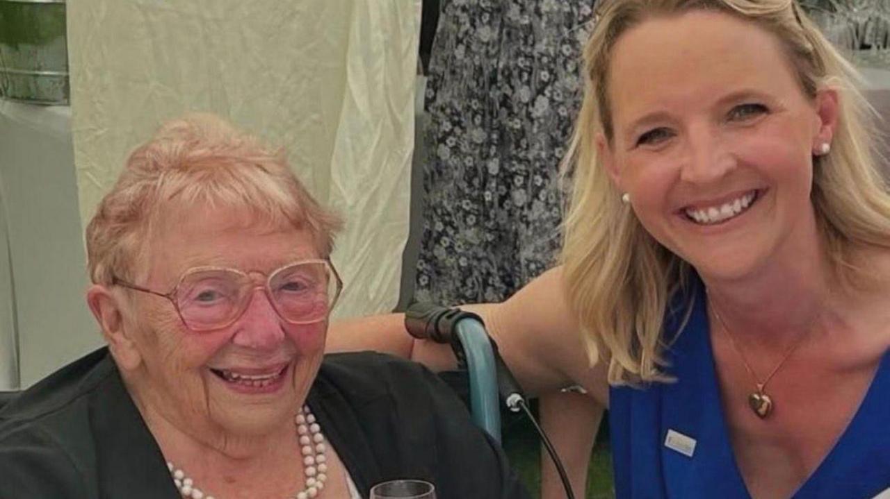 Rozelle Sutherland on the left and Kate Wright on the right. They are both smiling at the camera. Rozelle is wearing a black top with a pearl necklace and is wearing glasses. Kate is wearing a blue dress and is wearing a gold necklace.