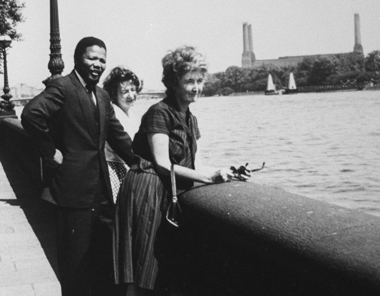 Nelson Mandela smiling in a black and white archive photograph between two women standing on the banks of the river Thames with Battersea power station in the background