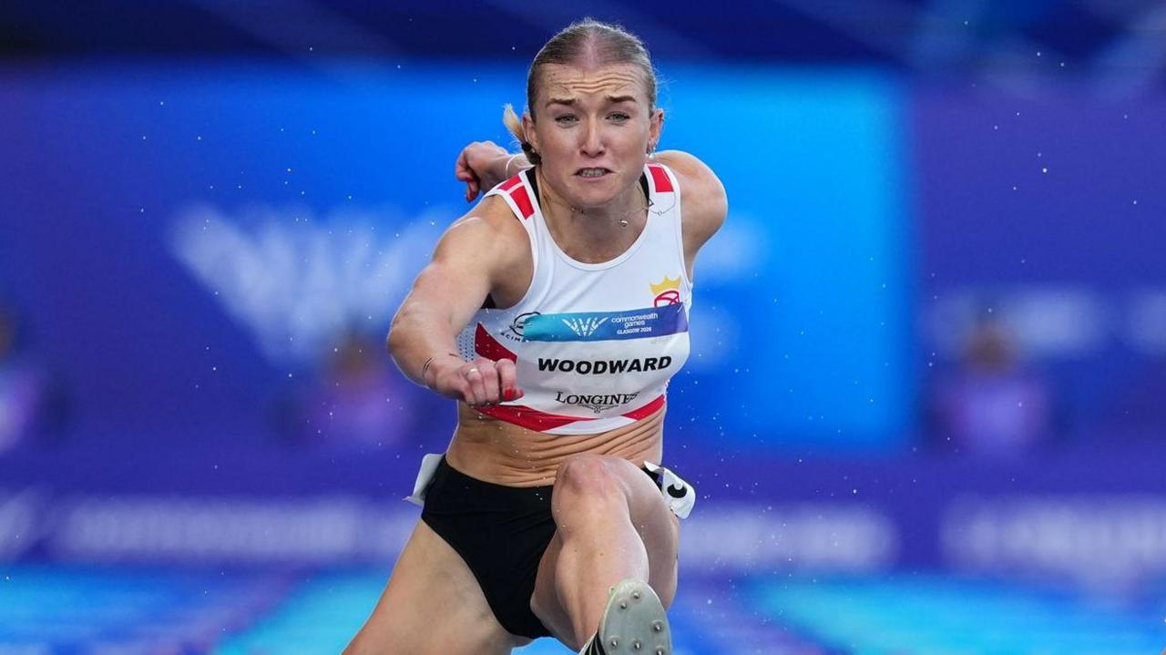 A head on image of Lucy Woodward, wearing a white vest with red trim and black shorts, jumping a hurdle