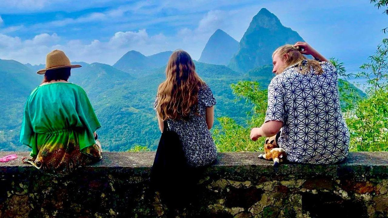 Two young women and a young man, all dressed in casual holiday clothes, sit on a wall with their backs to the camera, looking towards dramatically tapered mountains on the island of Saint Lucia. The landscape is covered in greenery beneath a blue sky.