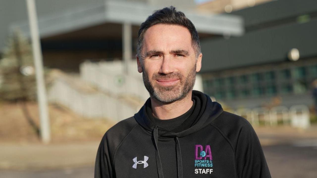 Alan Millar, a man with black hair and a short beard. He is standing outside a college campus building and is wearing a Dundee and Angus College hooded top.