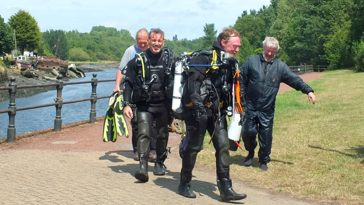 Two divers in full gear walking along the river bank after getting out of the River Wear. They are smiling. Two men are walking beside them and carrying equipment. Gary Bankhead is one of the divers. He is smiling and has short grey hair.

