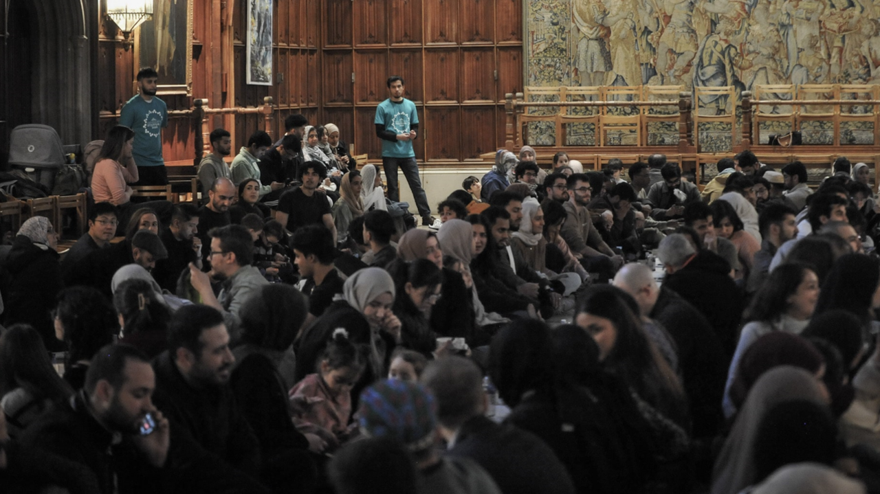 Lines of people sitting on the floor of a large room. They appear to be talking. Some women are wearing headscarves. A man in the foreground is talking on a mobile phone. Two men wearing green t-shirts are standing at the back of the room.