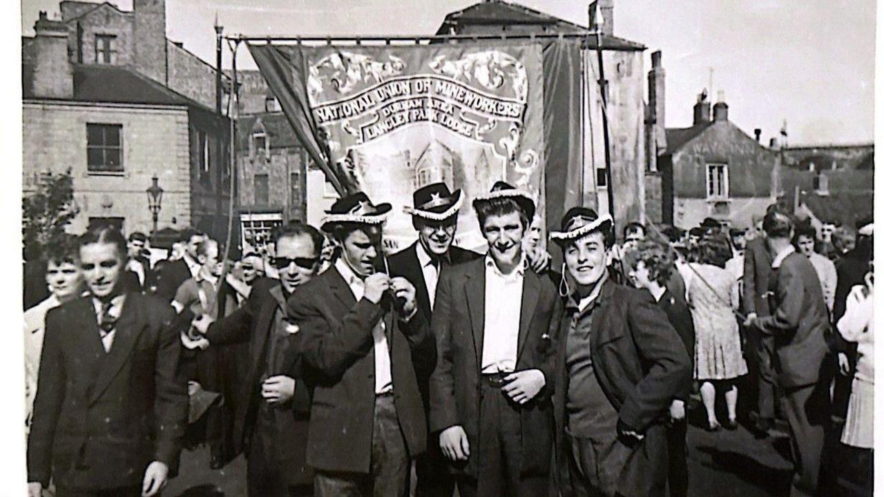 A black and white postcard of the Langley Park miners' banner. Lots of people in black blazers pose in front of the banner which is being held up by a mass of people. 