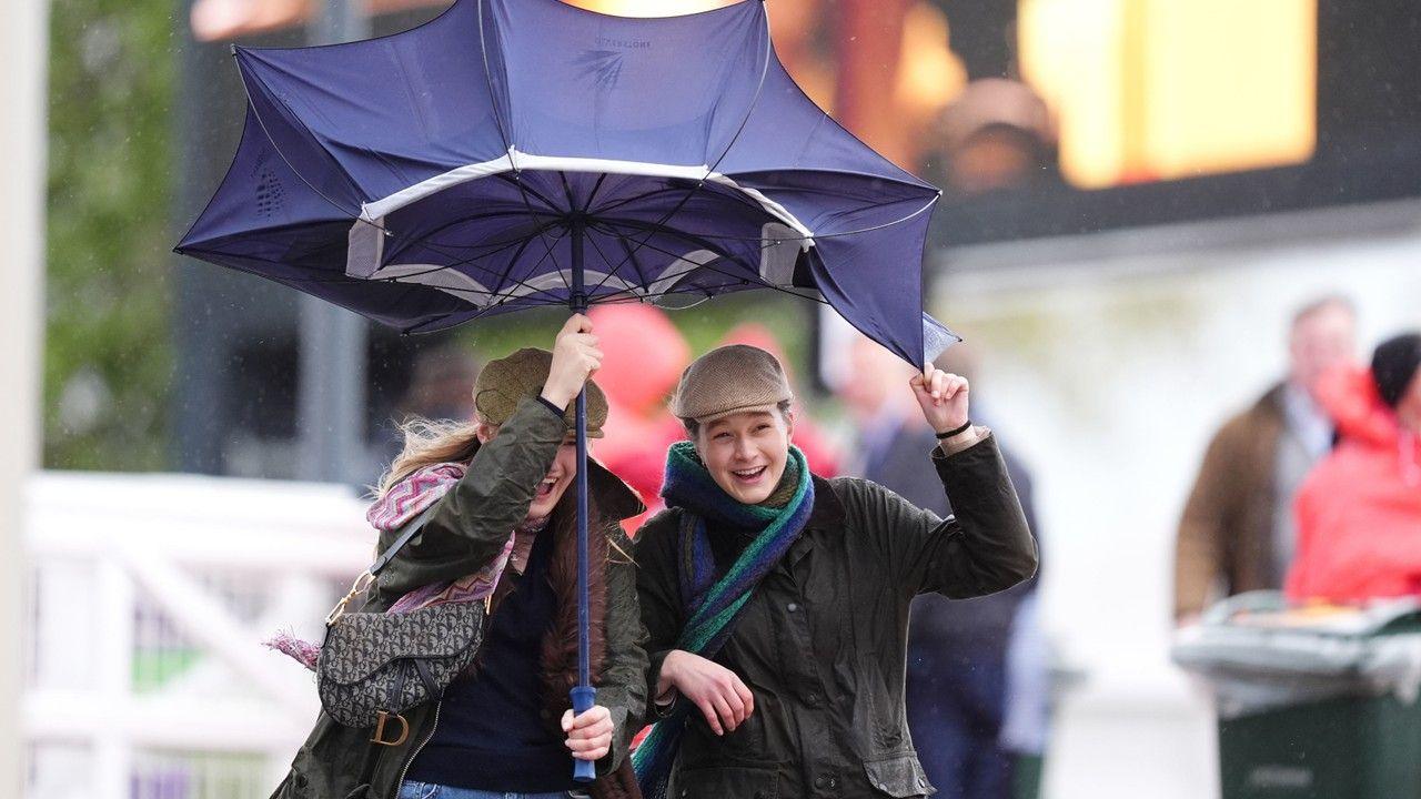 two young people wearing flatcaps and scarves, smiling whilst sharing a large umbrella that has blown inside out