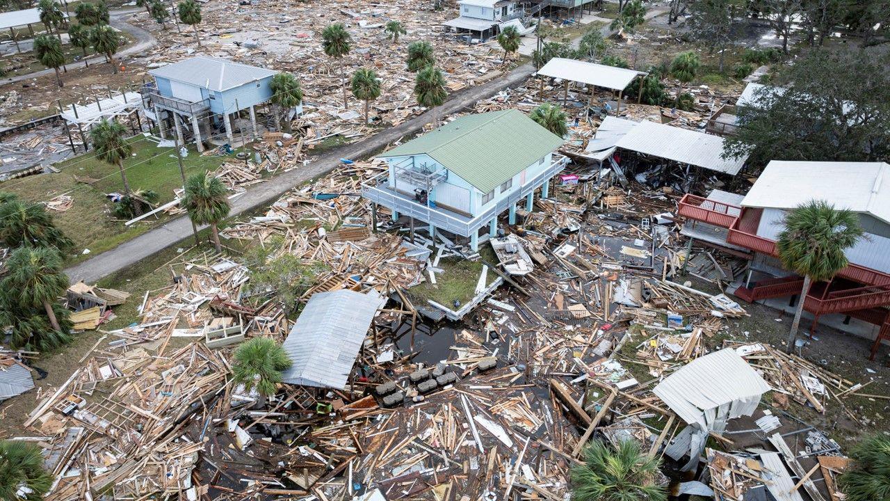A drone view shows flooded and damaged homes with pieces of wood covering a large, now sparse area following a hurricane