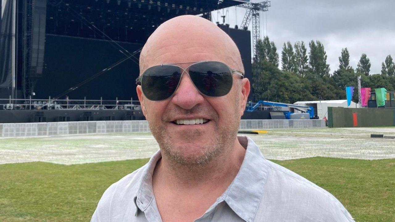 Alan Simms wearing sunglasses and smiling at the camera, he is wearing a grey shirt. Behind him is a large stage with a sign reading 'EMERGE' above it. There are a number of trees to the right of the stage, and a tree to the left of the stage. The sky is overcast. There are a number of people in the background, some of whom are wearing high-vis jackets.