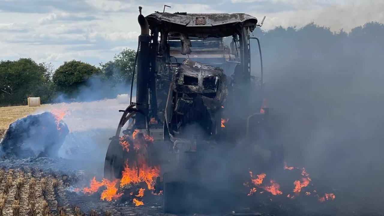 A blackened and burning tractor in a field. Its structure has warped in the fire, which is still blazing around its tyres with smoke rising. Behind is yellow stubble, two round straw bales can be seen, trees and blue sky.