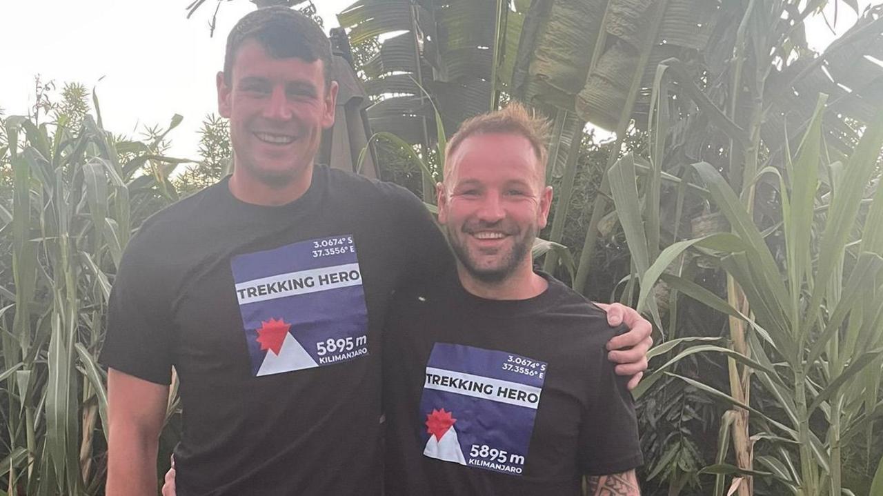 Rory Honeychurch and Ben Horn stood together wearing black t-shirts with the words 'Trekking Hero' on the front. They are both smiling at the camera. Behind them is long grass.