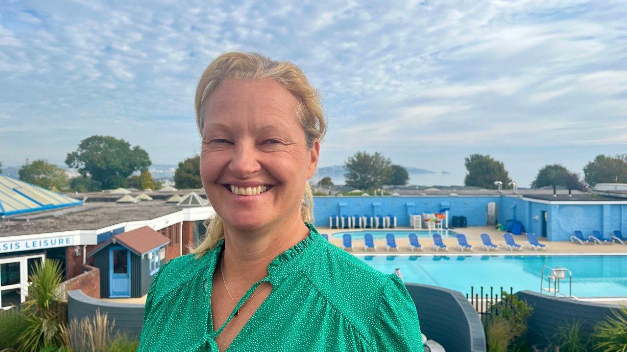 Claire Flower, owner of Beverley Park in Paignton, wearing a green and white top with her blonde hair tied back. Behind her you can see the swimming pool and beyond that, the sea at Torquay with mist on top.