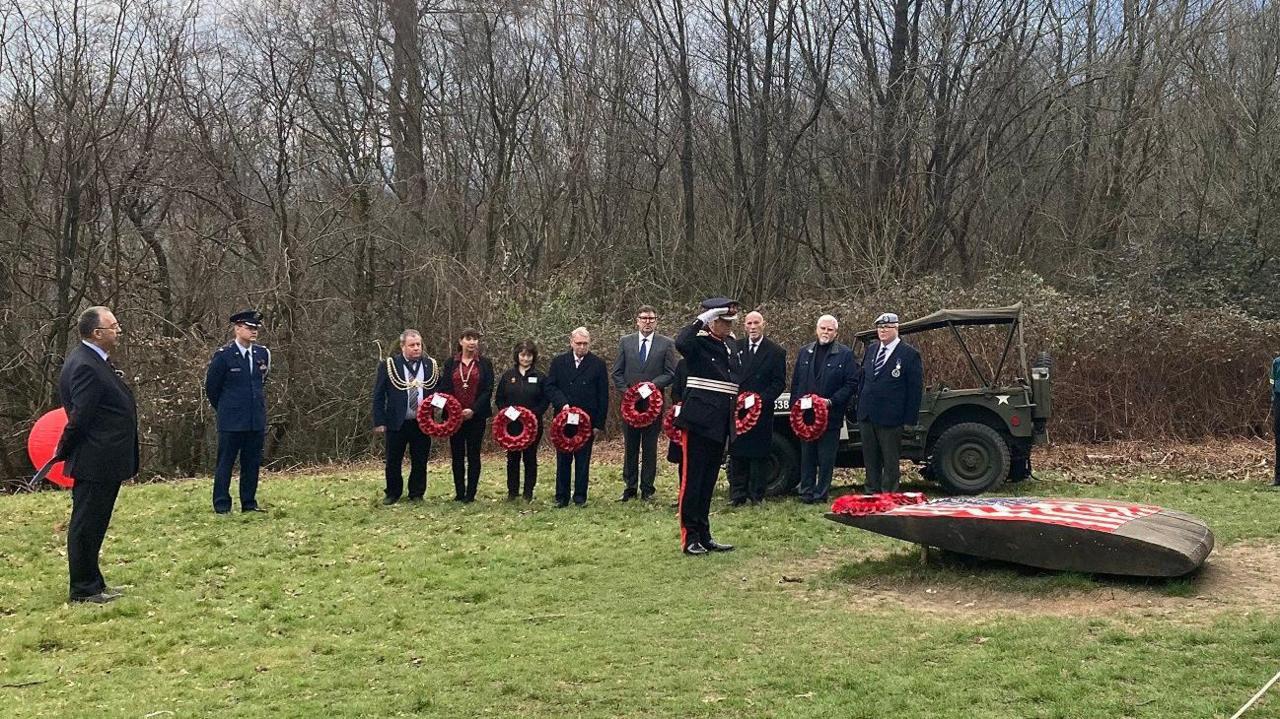 A group of people, some in military uniform and some holding wreaths, standing on a patch of grass before some trees and shrubs. One man is saluting a low wooden memorial in the shape of a plane wing tip. 