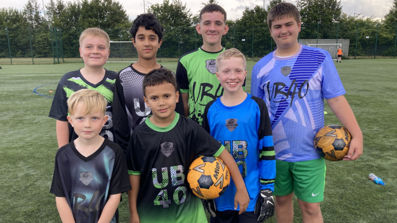 Seven young boys of different ages stand on the pitch wearing the kit. They are wearing blue tops, black and green tops and green shorts. Two boys are holding footballs.