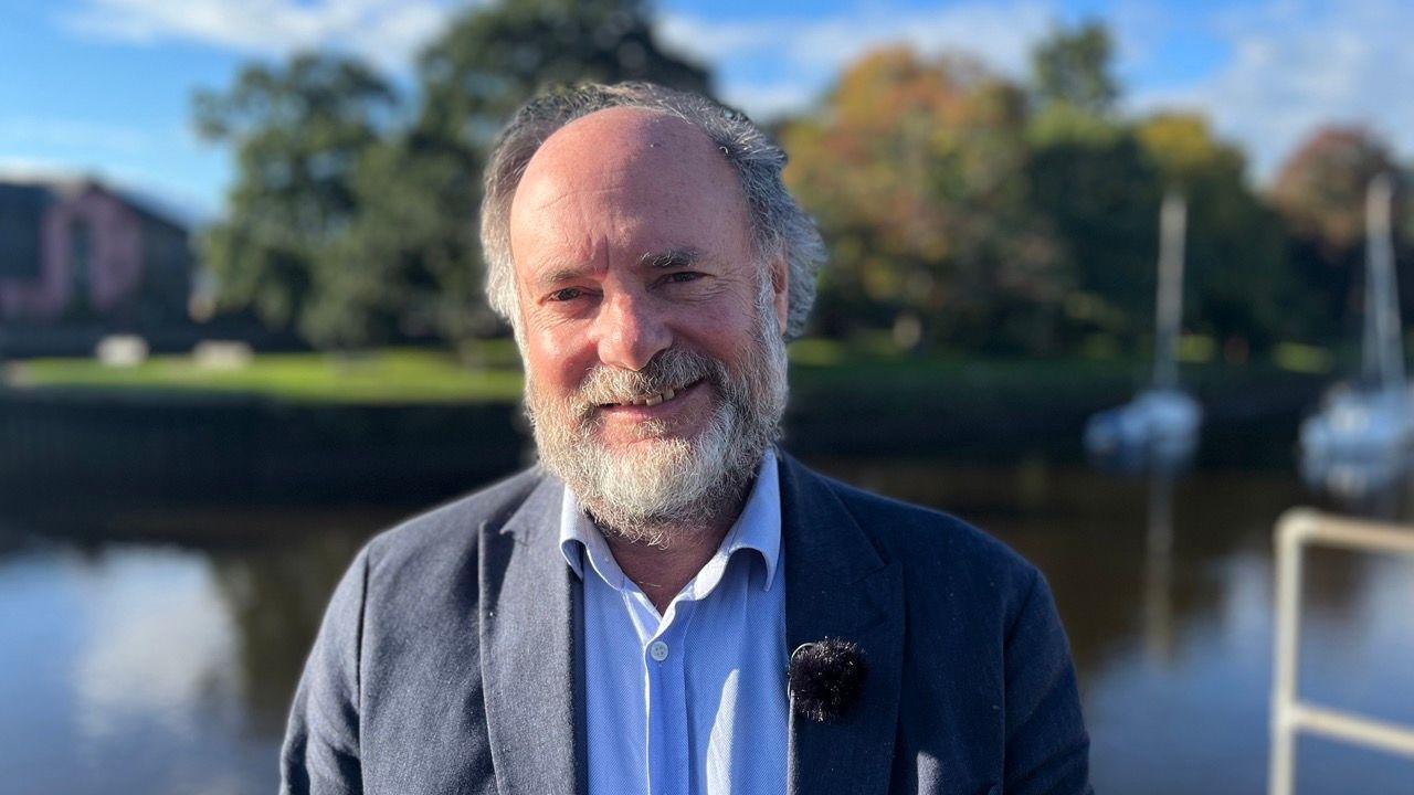 The leader of South Hams District Council, Julian Brazil, wearing a blue shirt and navy blue blazer, by the river in Totnes with a blurred background.