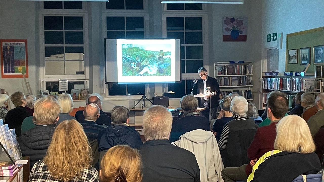 A room full of people listening to a speaker at the front with a presentation screen. An image is being projected on the screen - there are books on shelves behind the speaker.