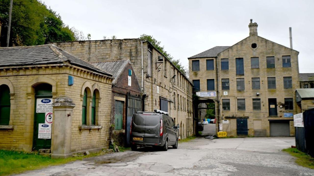A mill building which has a car park in front with a grey van parked there. 