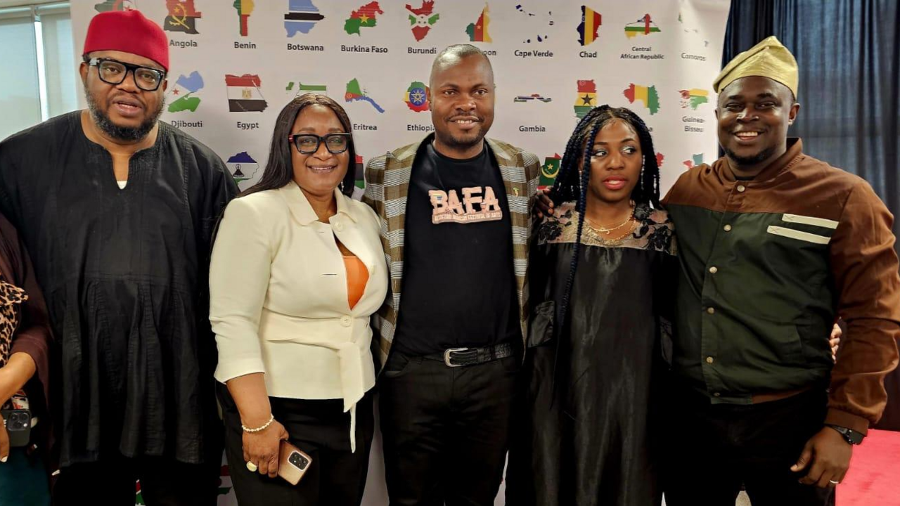 A group of six people, all from an African-Caribbean background, stand smiling in front of a display board which has the names and flag style maps of various African countries on it.