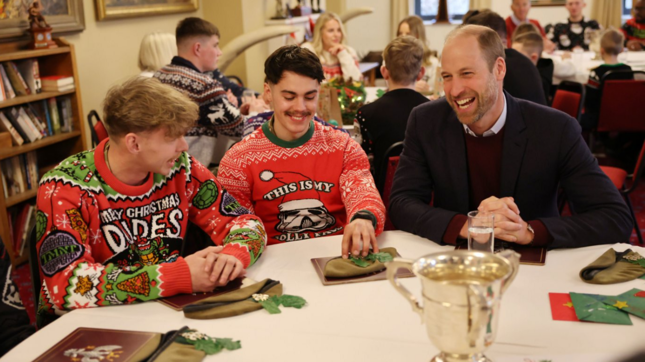 Prince William sitting at a table with two young military men wearing Christmas jumpers and smiling, in a room with lots of other families 