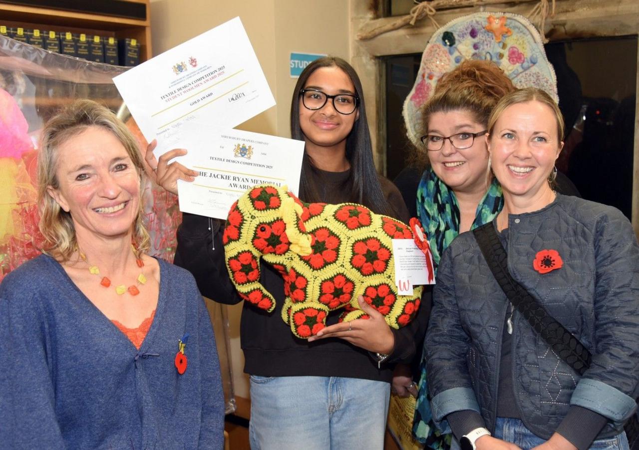 A winnning student with long black hair and glasses holds a yellow and red woollen elephant in one hand and certificates in the other. She is flanked by three women and surrounded by other textiles work.