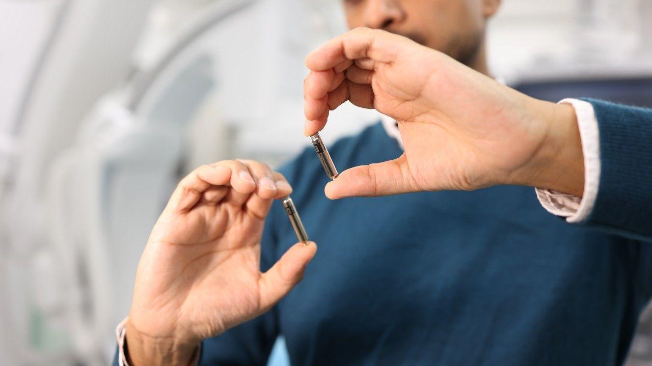 Hands hold up two small metal cylinder shaped pacemaker devices which are smaller than a triple A battery. These will be inserted directly into two chambers of the heart.