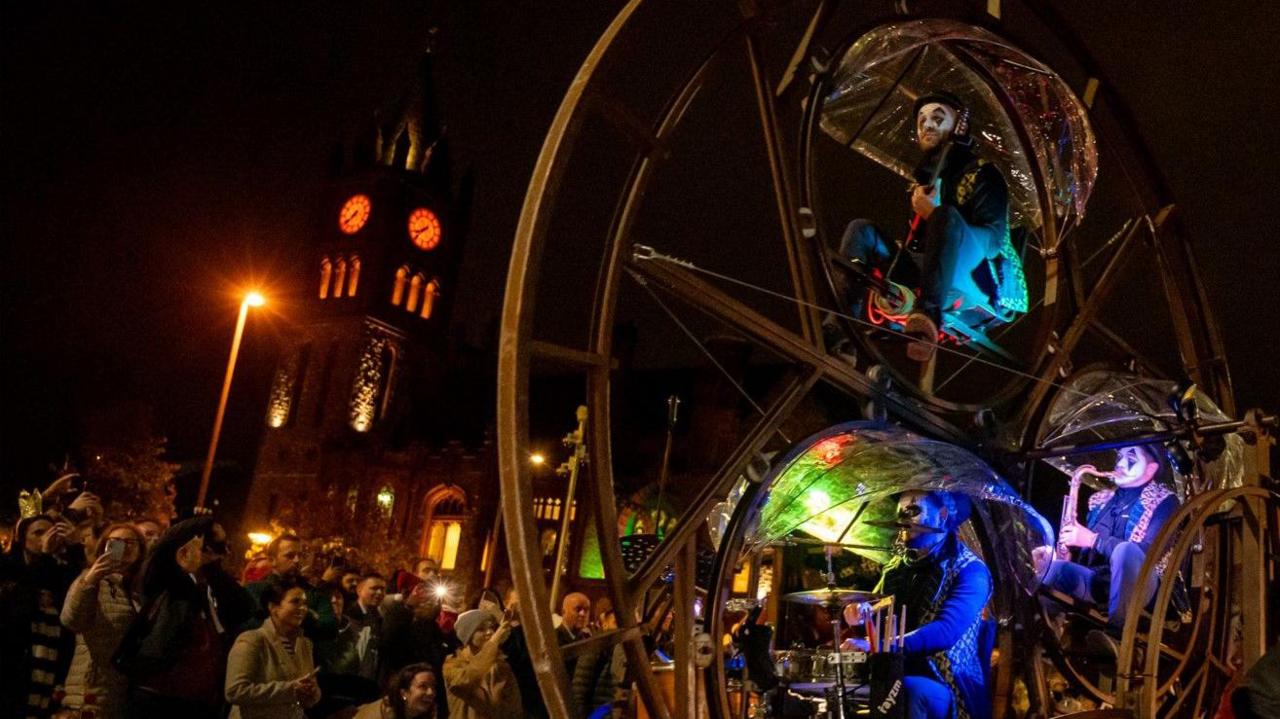 Crowds watching musicians on a large wheel structure near Guildhall Square 