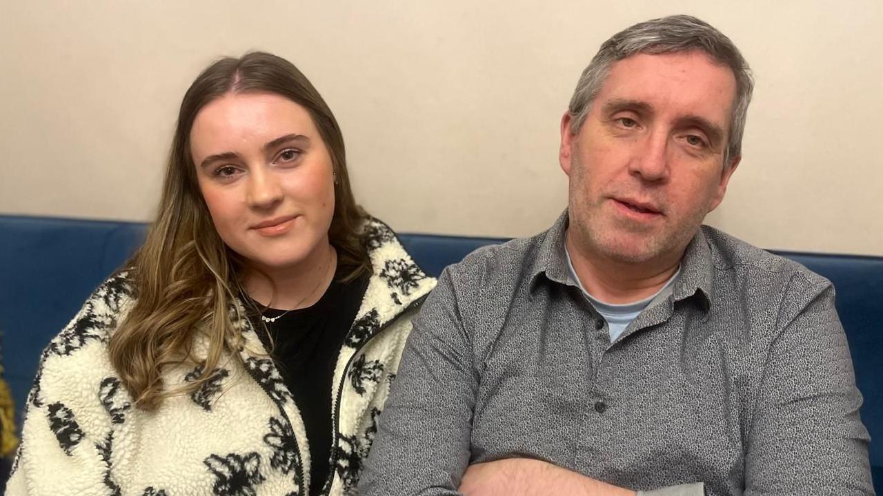 Aimee sitting on a sofa with her dad Damien. Aimee has long brown hair with blonde ends, she's wearing a black top and a cream zip up cardigan with black flowers. Damien has light grey short hair and is wearing a grey shirt with a light blue top underneath.