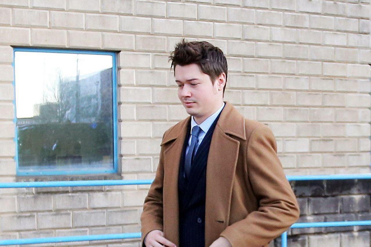 A man in a light brown overcoat with a navy suit jacket, blue tie and light blue shirt underneath. He has short brown hair and has a slight grin. Behind him is a wall of grey bricks with a window.