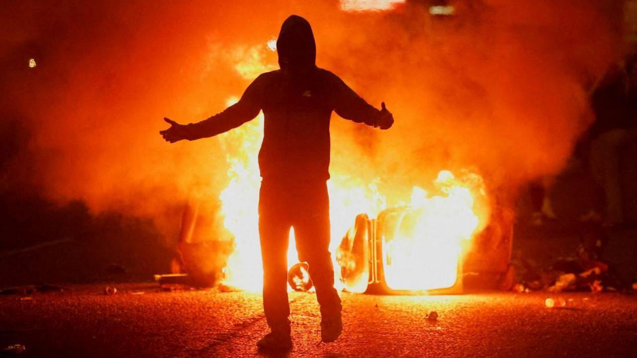 A man wearing dark clothing and with his hood pulled up stands silhouetted with his arms outstretched in front of a burning wheelie bin. Flames are billowing from the bin.