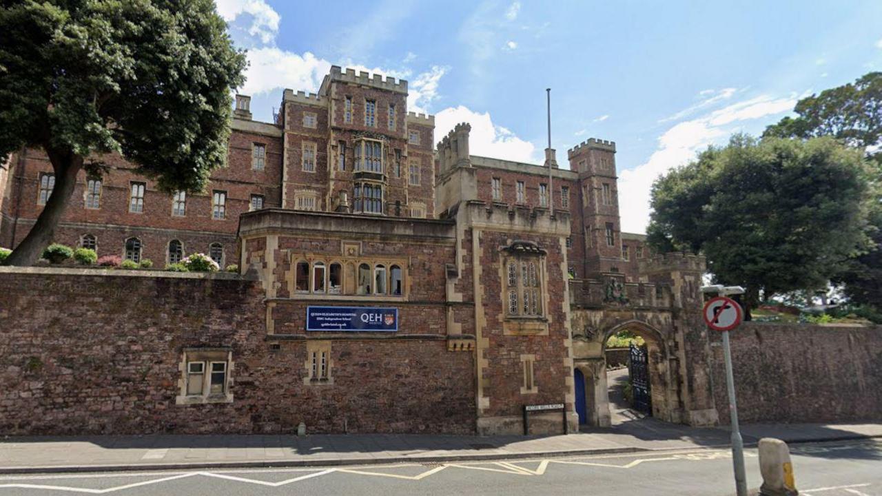 A screenshot from google street view showing the Queen Elizabeth’s Hospital arched entrance beside the road. It is a large 14th century brick building, with castle-like turrets, lots of windows and a flagpole. There is a brick wall surrounding the perimeter and a gate house building. There is a blue sign on the wall which reads Queen Elizabeth’s Hospital. 