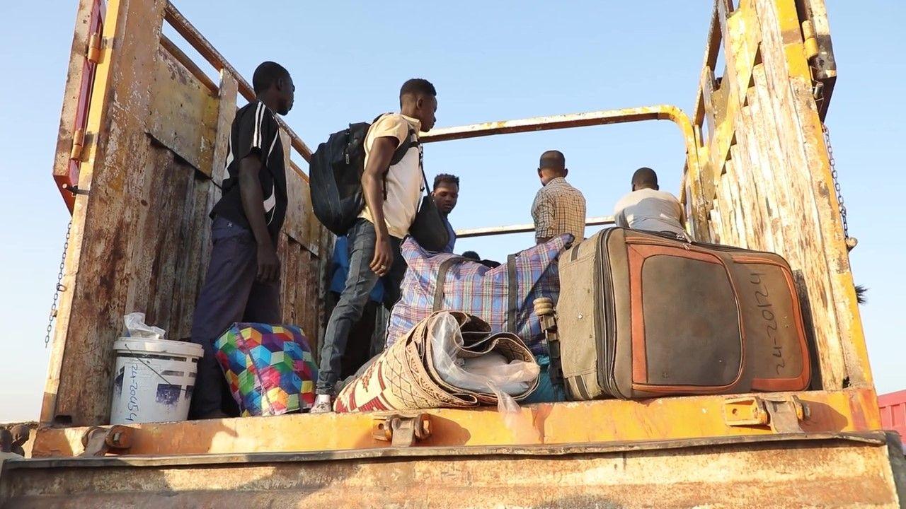 A group of men are standing in the back of an open lorry, a suitcase, a rolled-up carpet and some other luggage can be seen in the foreground at the edge of the back of the vehicle.