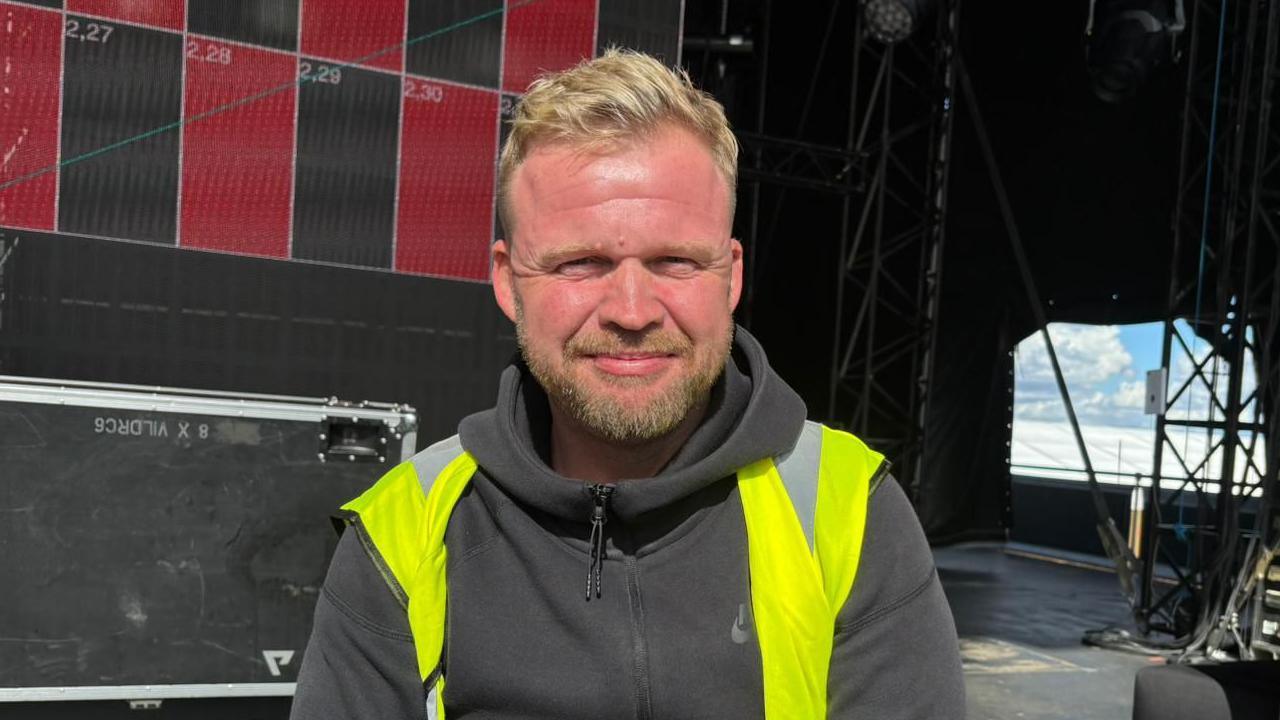 Chris Halls wearing a hi-vis jacket and a black hoodie. He is sitting on the main stage and smiling at the camera. He has blond hair and stubble.