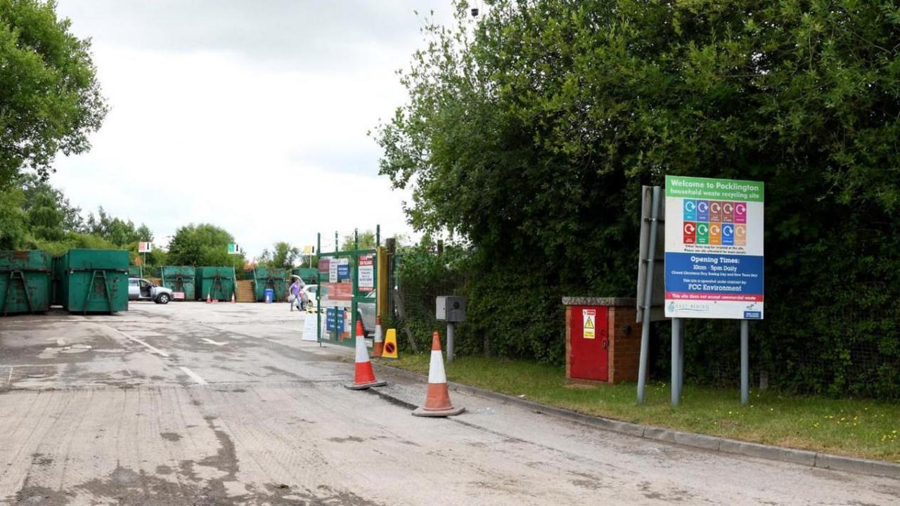 The entrance to Pocklington household waste recycling centre, looking towards green skips for holding waste. In the foreground is a sign, two cones and a large, green tree