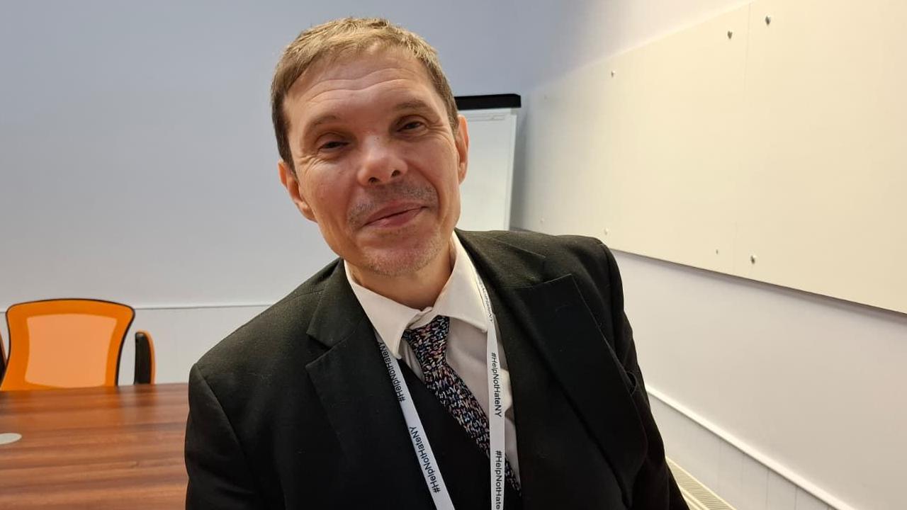 A man wearing a suit and tie and lanyard which reads "#Hope Not Hate NY" smiles at the camera. He is standing in a meeting room.