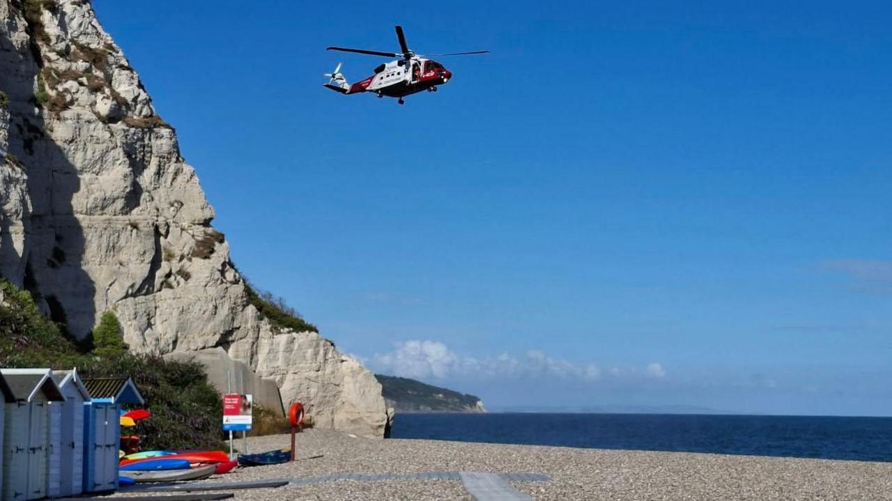 A rescue helicopter flying over a beach. There is a cliff face to the left. The sky is blue. The water is a dark blue. 