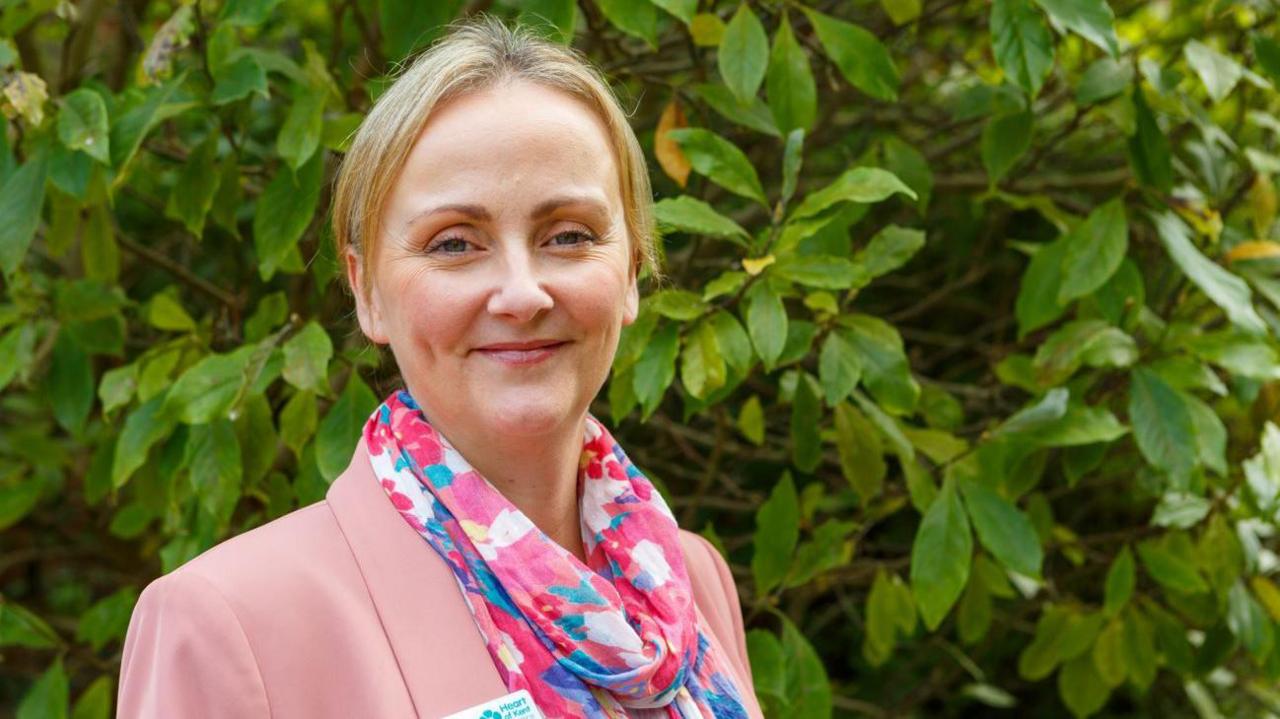 Rachel Street stands in front of a hedge. She is smiling and wearing a pink blazer and a multicoloured scarf. She has short blonde hair.