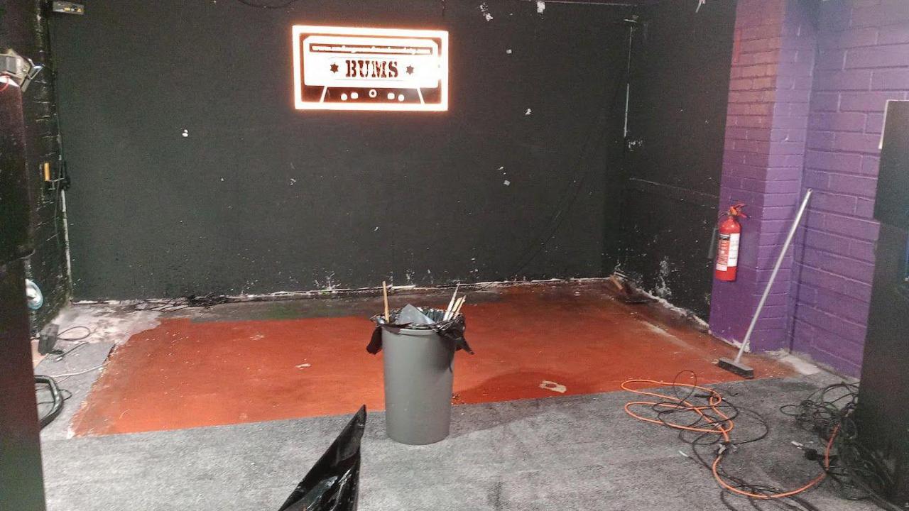 Water damage shown on the floor and around the wall inside the Barrow Undergound Music Society.  Bright logo in the shape of a cassette tape containing the words "BUMS" is shown on the wall.