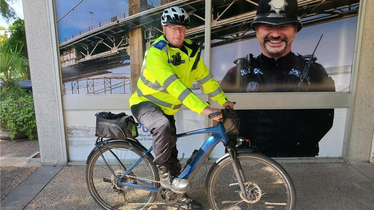 PCSO Paul Fudge in a florescent yellow jacket and white helmet, sitting on a blue e-bike